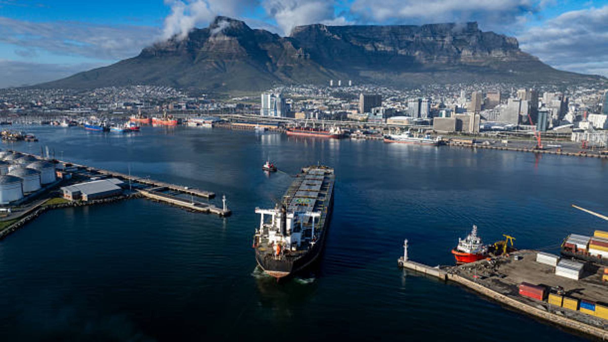 Cargo vessel moving through a working harbour, with port infrastructure and container activity along the quay
